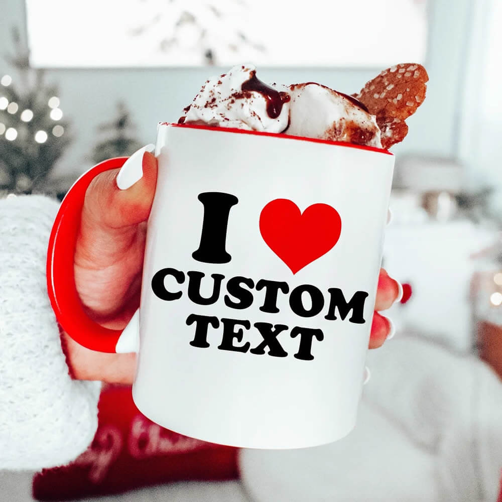 White mug with 'I ❤️ CUSTOM TEXT' text, held by a person with hot chocolate and marshmallows.