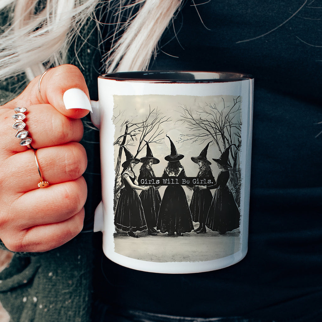 Mug with a design of women in hats and the text 'Girls Will Be Girls' held by a person.